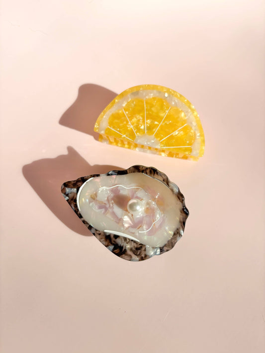 Oyster Claw Clip with Pearl by Line&Co displayed beside a lemon slice prop. Chic and playful hair accessory for beach-inspired fashion.