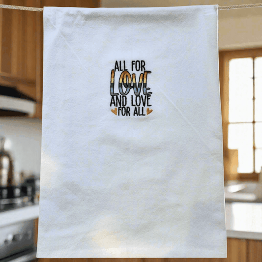 Soft flour sack kitchen towel hanging in a cozy kitchen, featuring rainbow embroidered 'All for Love and Love for All' message at the center.