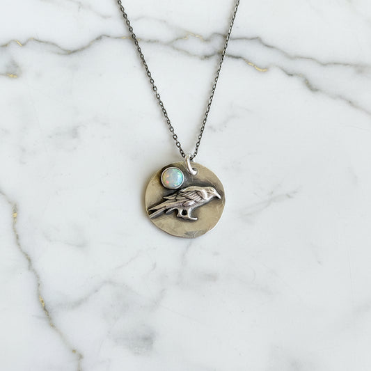 Silver necklace with a round pendant featuring a hand and opal stone on a textured gray surface.