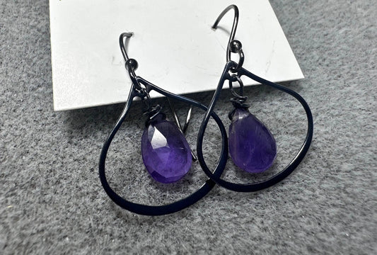 Amethyst gemstone earrings in oxidized silver Marrakesh-style hoops, showcasing a deep purple teardrop design on a gray fabric surface.