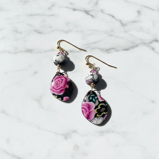 A pair of dangle earrings with gold-tone fishhook wires, small marbled beads, and teardrop-shaped charms featuring a pink rose and floral pattern on a black background, set against a plain white background.