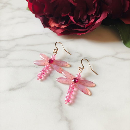 Pink dragonfly earrings with beaded wings and gemstone accents on marble surface