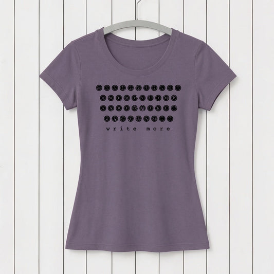 Lavender women's T-shirt made from soft organic cotton with black graphic print of a vintage typewriter keyboard and the words 'write more' at the bottom, hanging on a white wooden wall