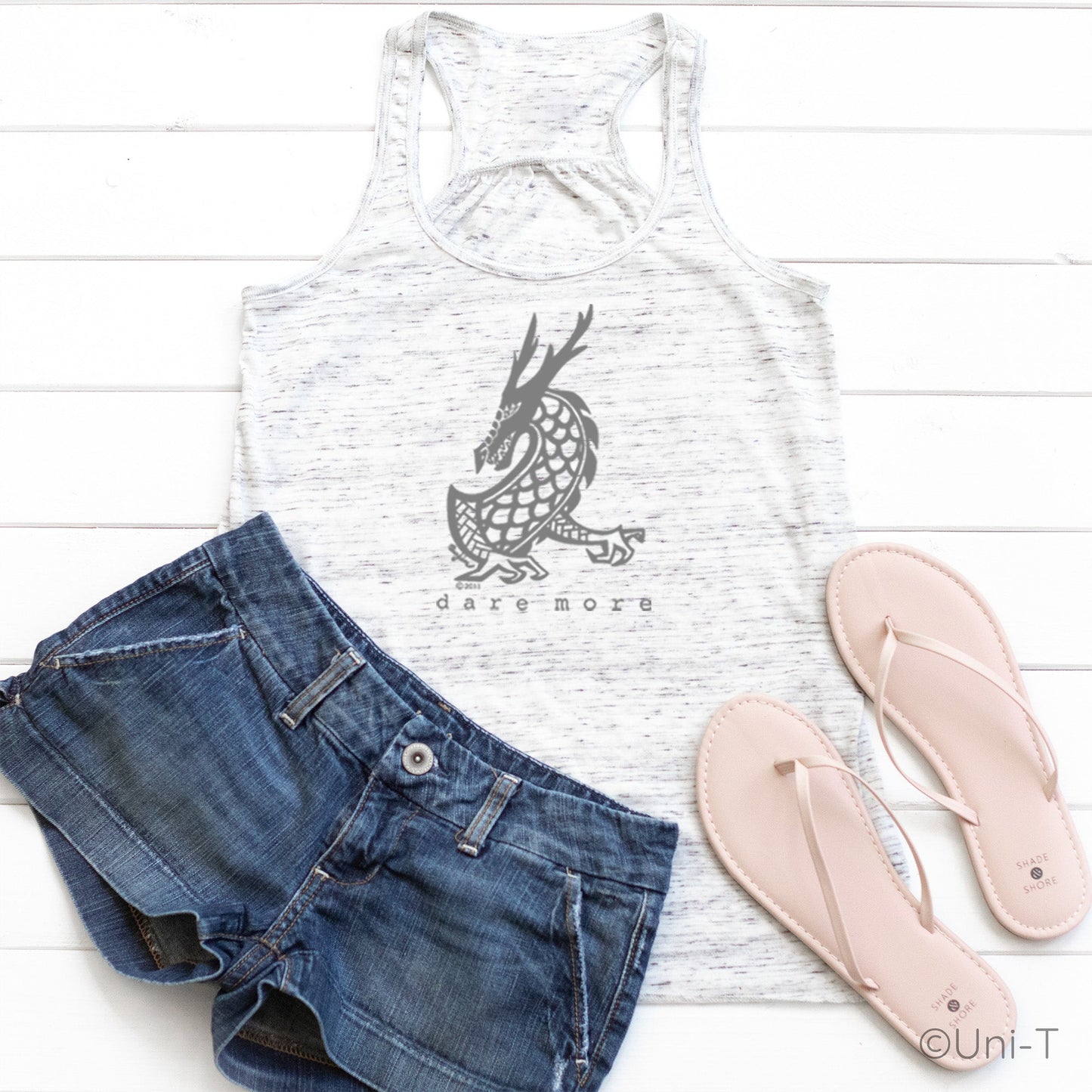 White speckled Dragon Flowy Tank Top Women styled with denim shorts and sandals. Features dragon design and 'dare more' text for bold living.