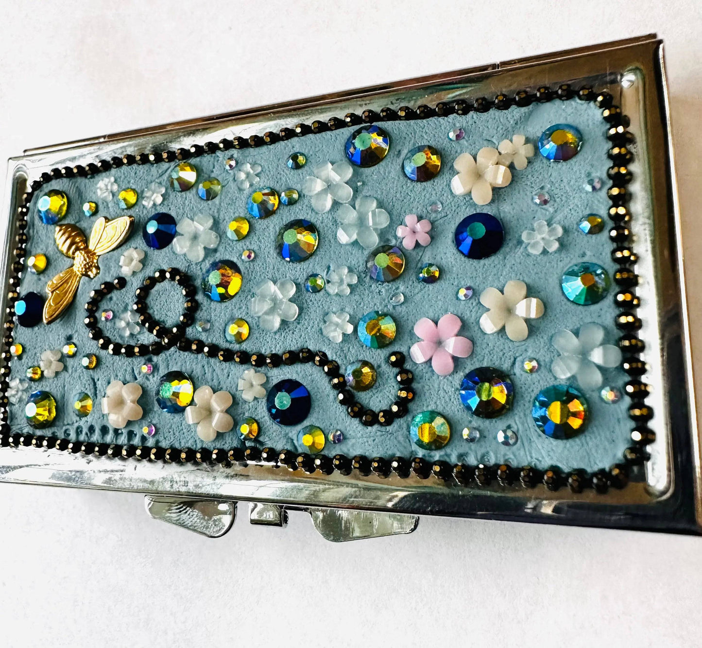 Decorative round brass pill box with blue background, floral accents, and sparkling rhinestones, designed for storing pills, jewelry, or makeup.