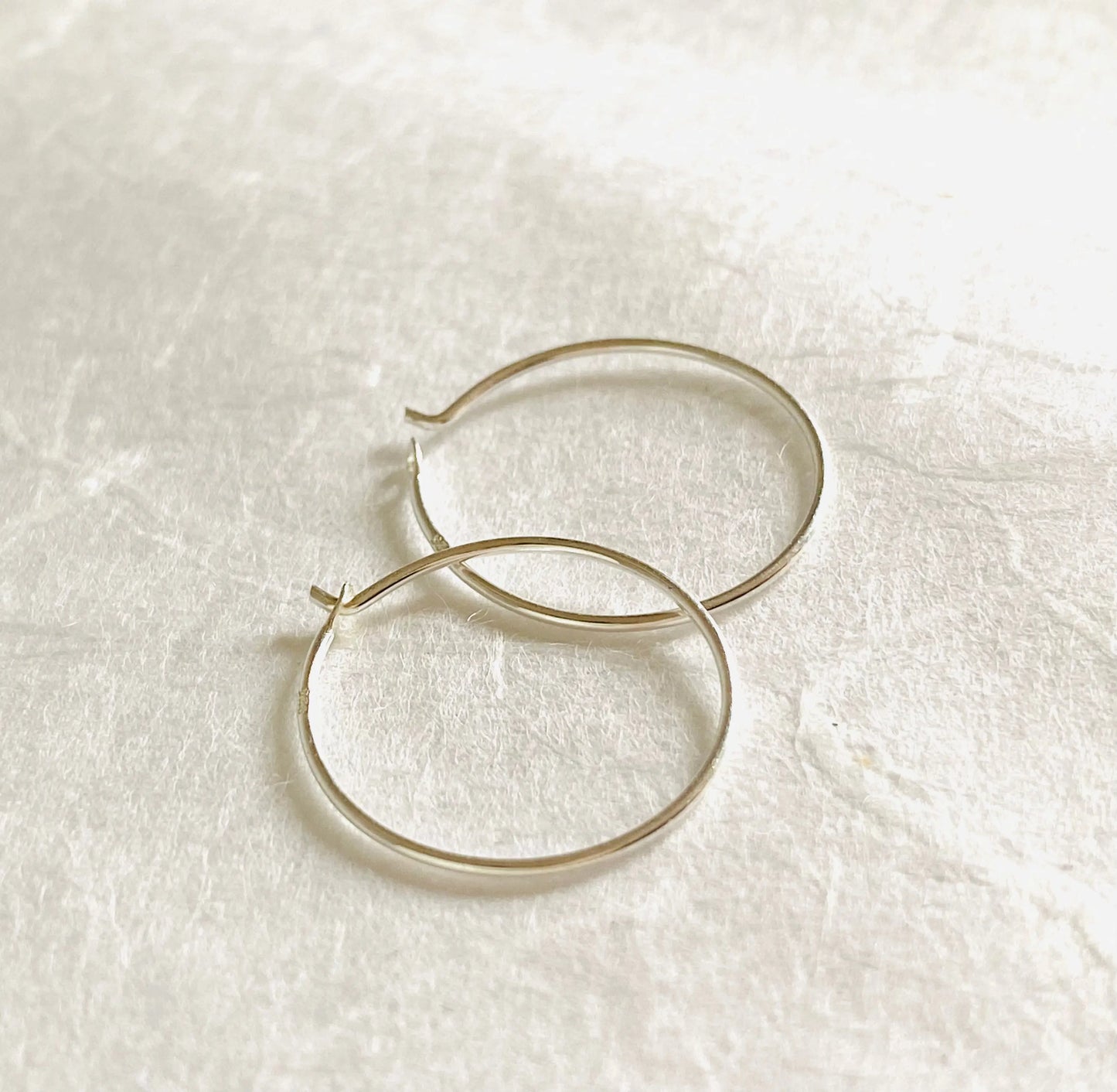 Minimalist 3/4” sterling silver hoop earrings on textured white surface. Elegant and lightweight hoops crafted from 22 gauge silver wire.