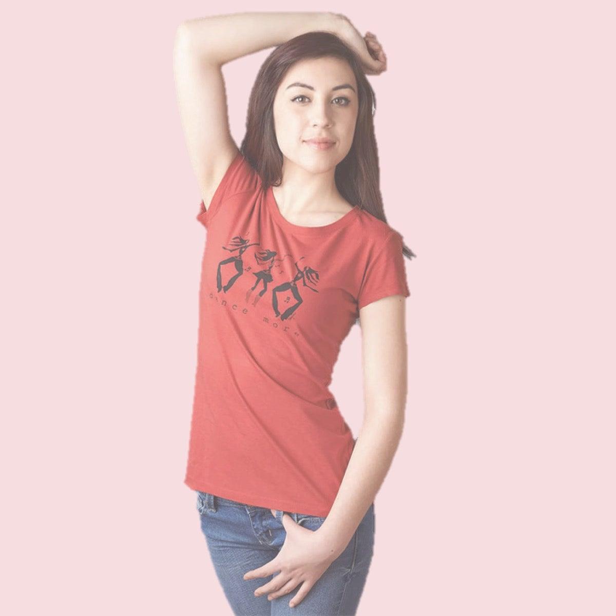 A woman with long brown hair poses with one arm raised, wearing a red t-shirt with a black abstract print and blue jeans, standing against a pale pink background.