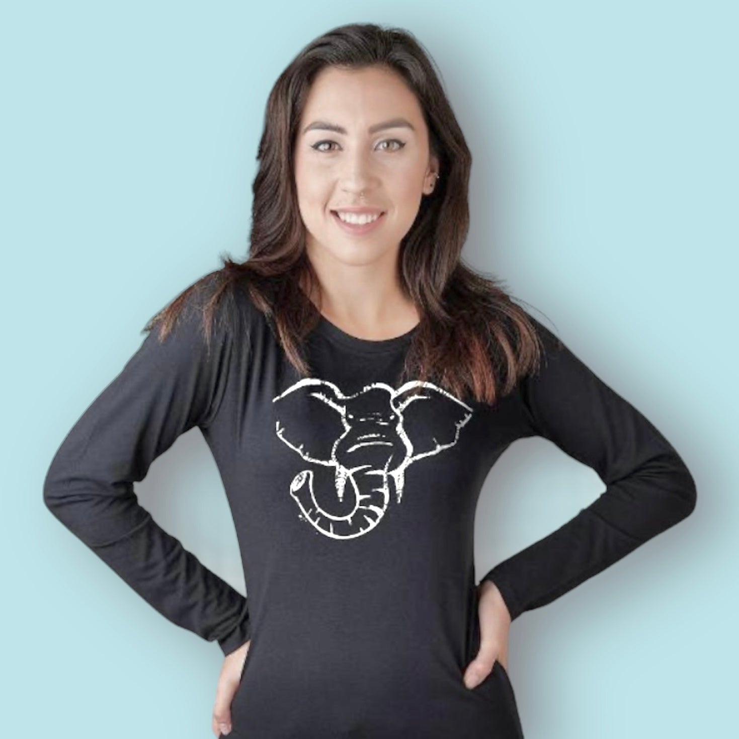 A woman with long dark hair smiles, wearing a black long-sleeve shirt featuring a white minimalist elephant design on the front, standing against a light blue background.