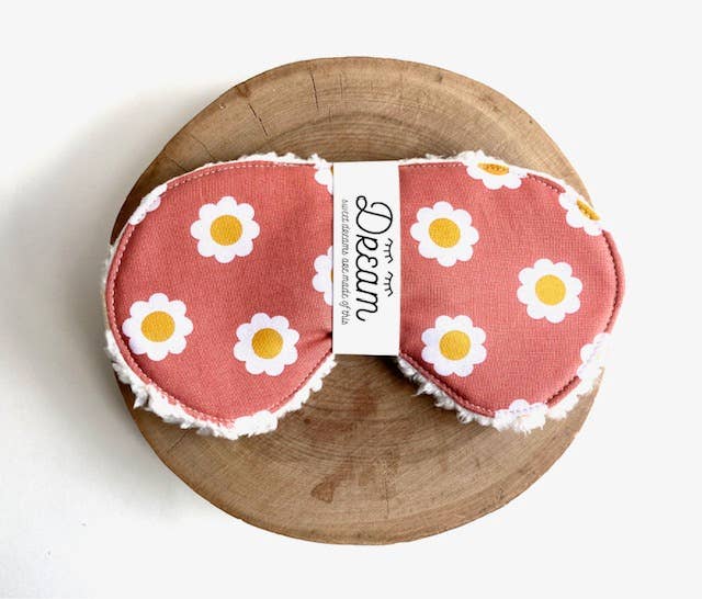 Cotton sleep mask with terra cotta daisy design shown on a round wooden board. Features soft Sherpa lining for enhanced relaxation.