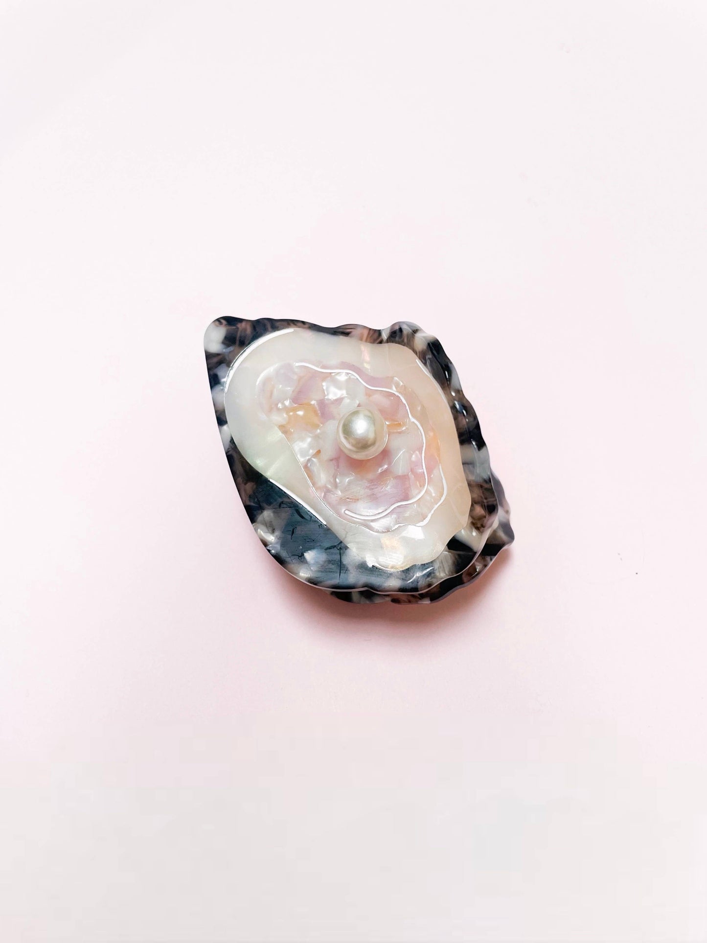 Close-up of Line&Co's Oyster Claw Clip with Pearl on soft pink surface. Features shimmering shell texture and central pearl accent.