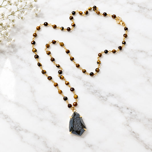 A beaded necklace with brown and black tiger's eye beads and a dark gray stone pendant, laid out on a white surface.