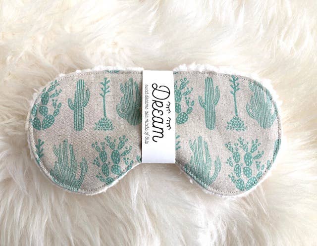 Sleep Mask Saguaro with green cactus print on beige cotton, laid on a fluffy surface to highlight its Sherpa backing and comfort.
