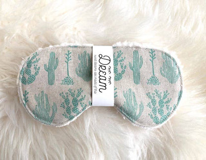 Sleep Mask Saguaro with green cactus print on beige cotton, laid on a fluffy surface to highlight its Sherpa backing and comfort.
