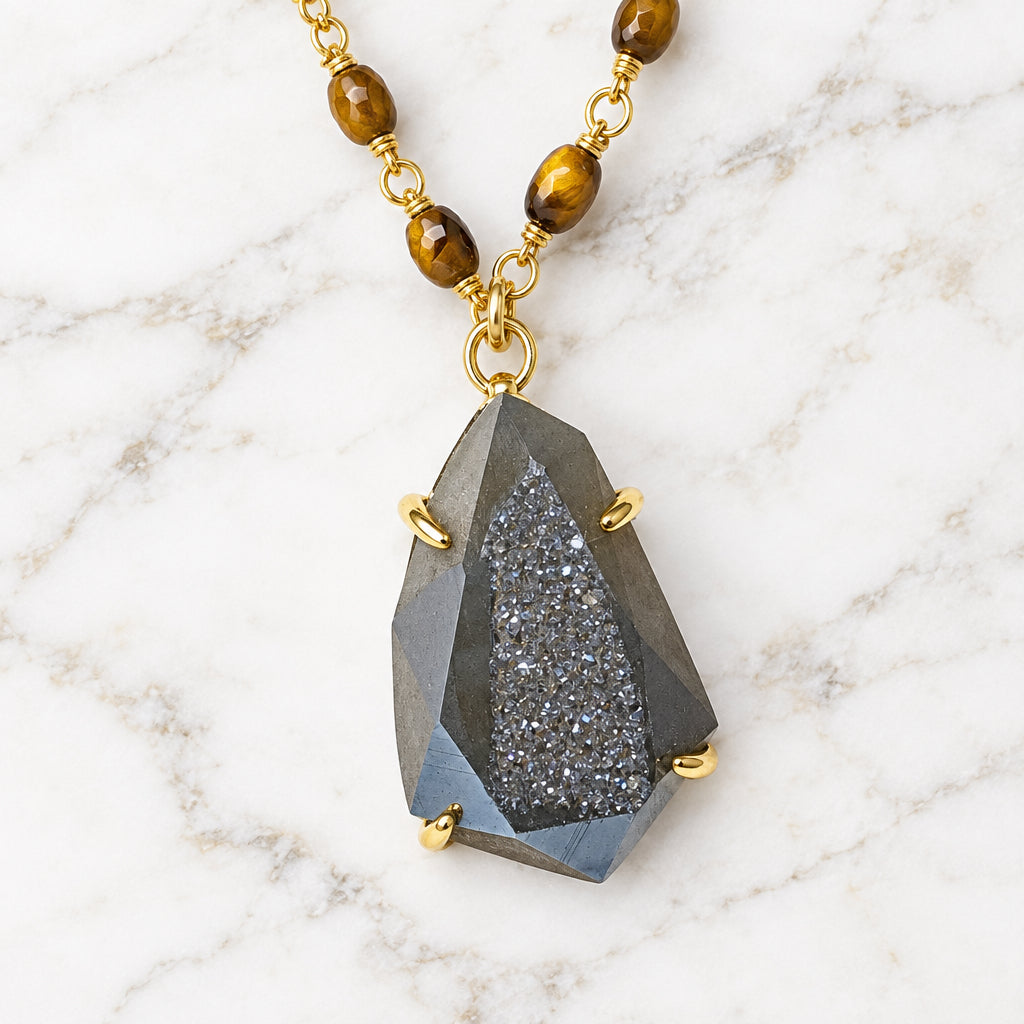 A faceted dark gray crystal pendant necklace with gold-toned metal settings and a gold beaded chain, displayed on a white surface next to a textured tree branch.