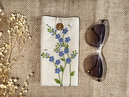 Embroidered linen glasses case with blue forget-me-not flowers and bees on cream fabric, padded for scratch-free eyewear storage.