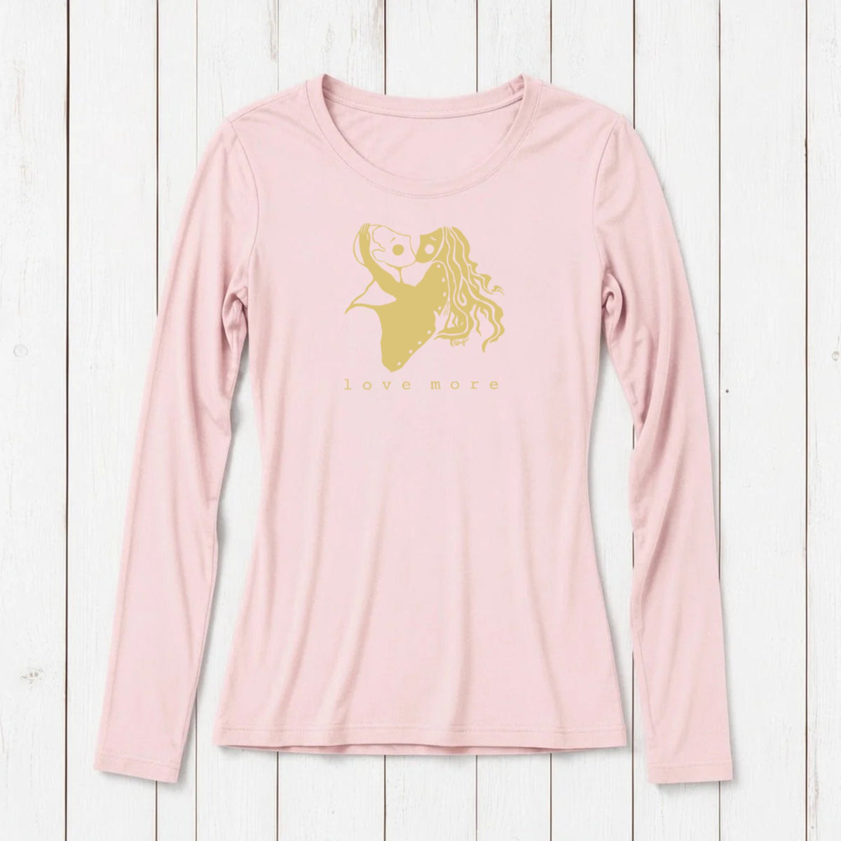 Light pink long sleeve T-shirt with a romantic gold graphic of a girl holding a heart-shaped balloon, featuring the words love more, on white wooden background
