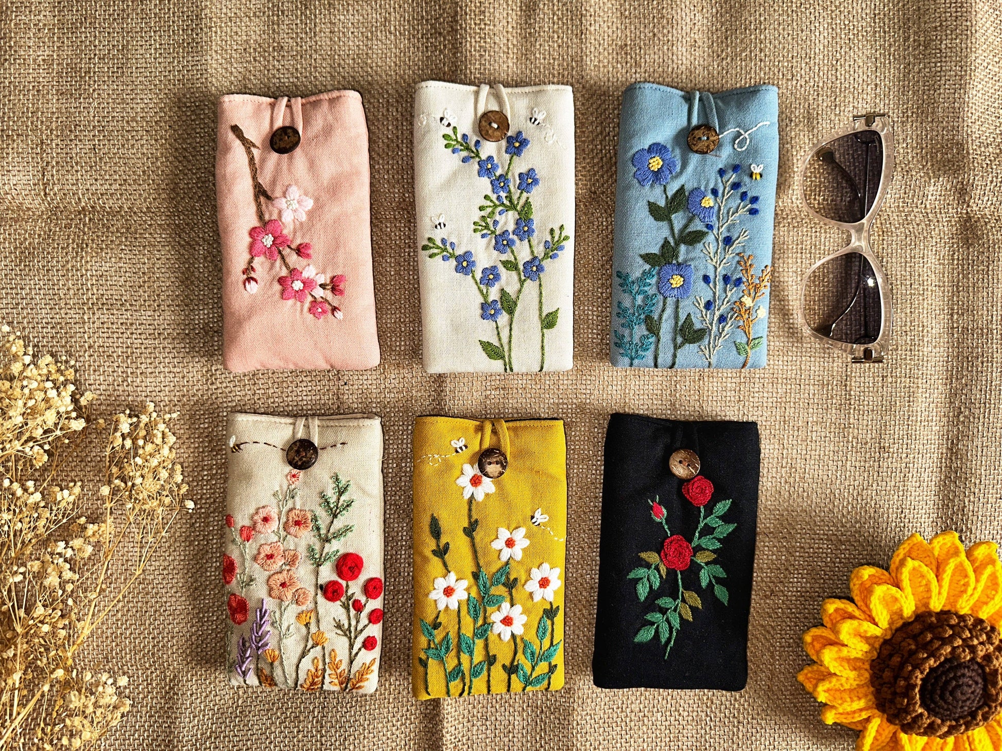 Set of six embroidered linen glasses cases with floral designs and button closures, offering stylish padded protection for eyewear.