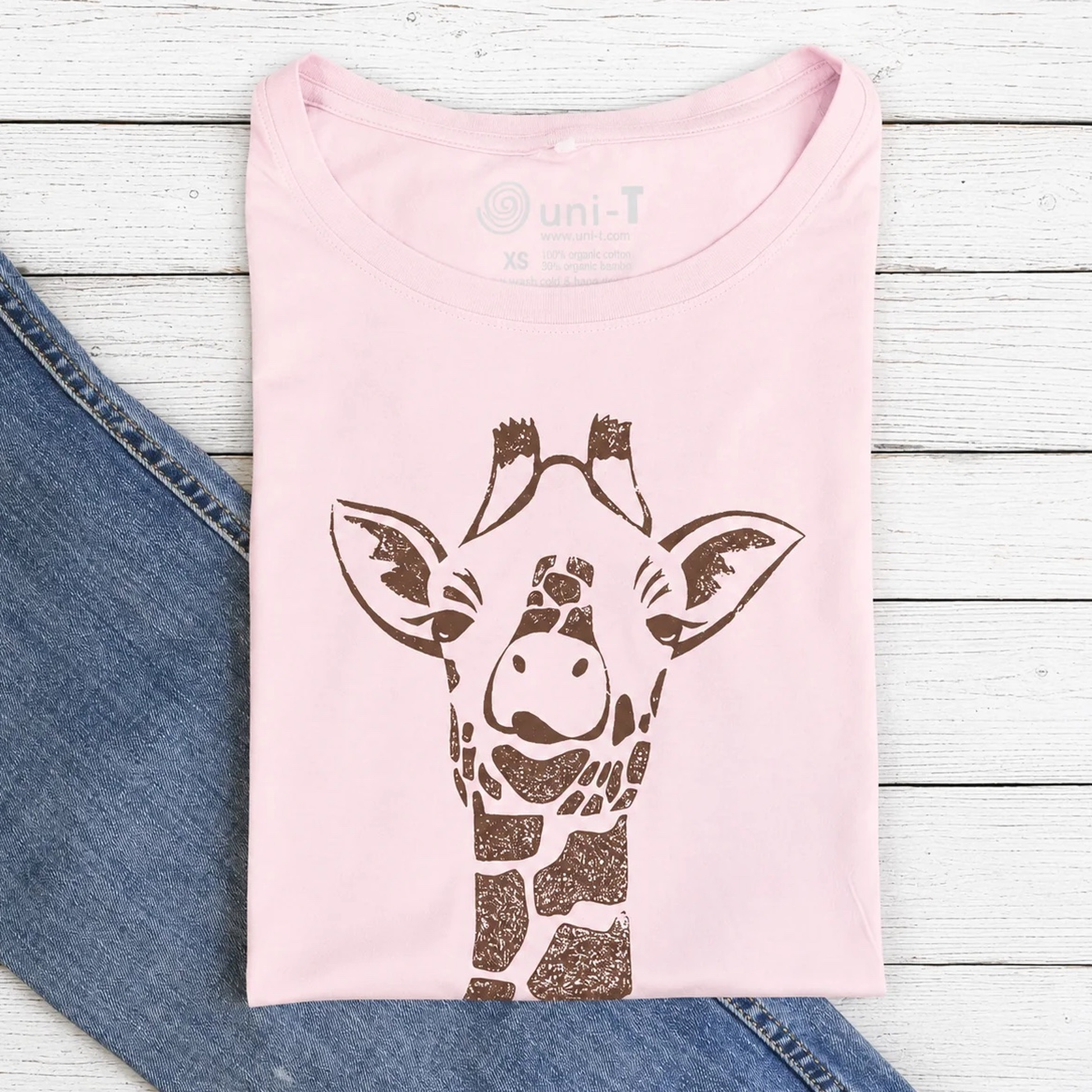 Light pink women's T-shirt featuring a brown giraffe graphic design, made from sustainable bamboo and organic cotton, laid on white wooden background with denim jeans partially visible