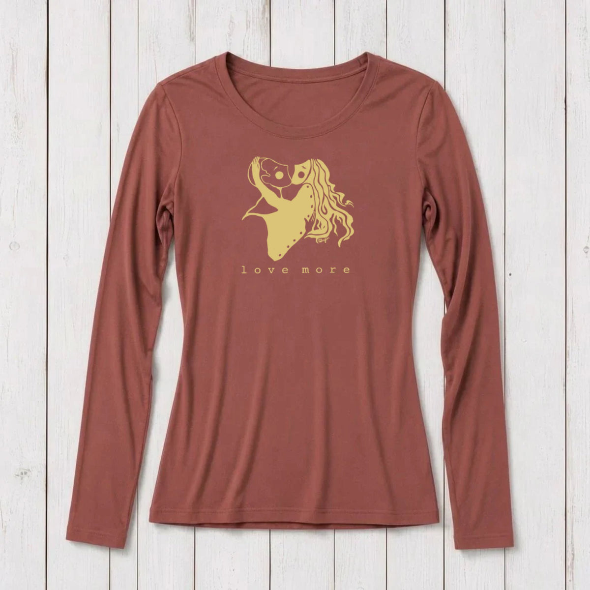 Long sleeve women's graphic t-shirt in rust color with a romantic heart and girl illustration, featuring the words love more, on a white wooden background