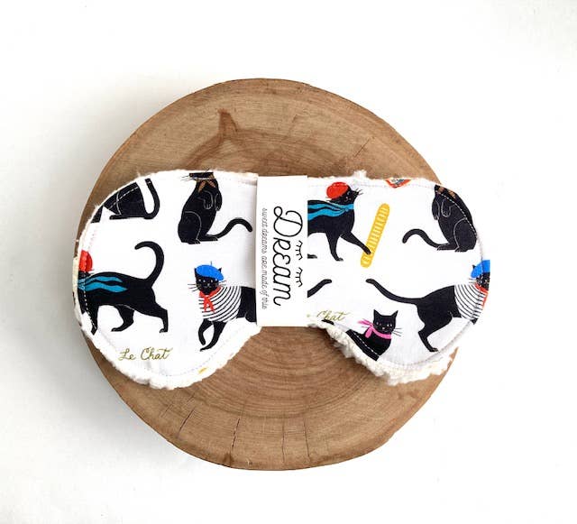 Le Chat Cotton Sleep Mask displayed on wooden base, showcasing black cat pattern and Sherpa lining for cozy, relaxing sleep support.
