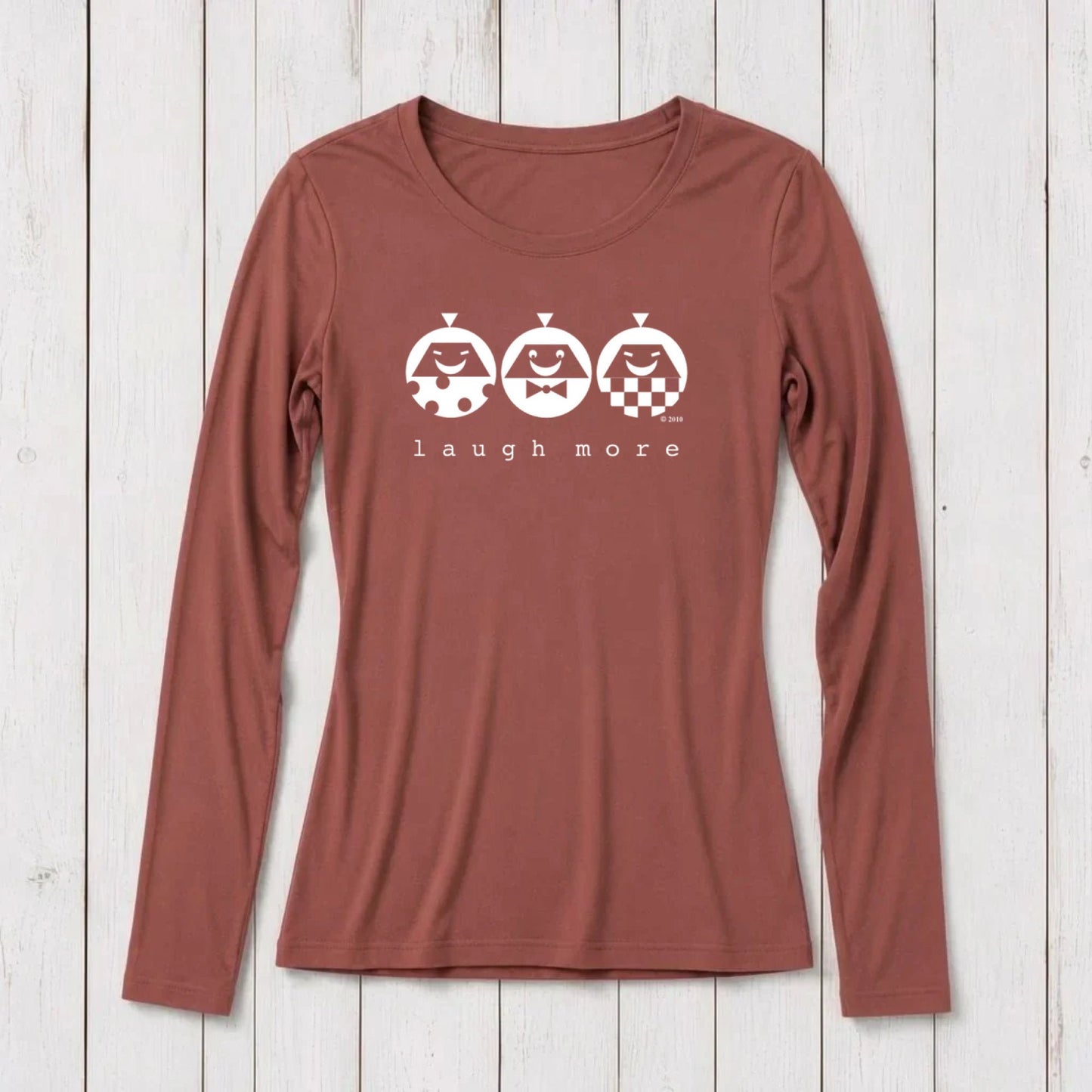 Rust-colored long sleeve T-shirt for women with white graphic design of three smiling faces with different patterns and the text 'laugh more' underneath, made from bamboo and organic cotton.