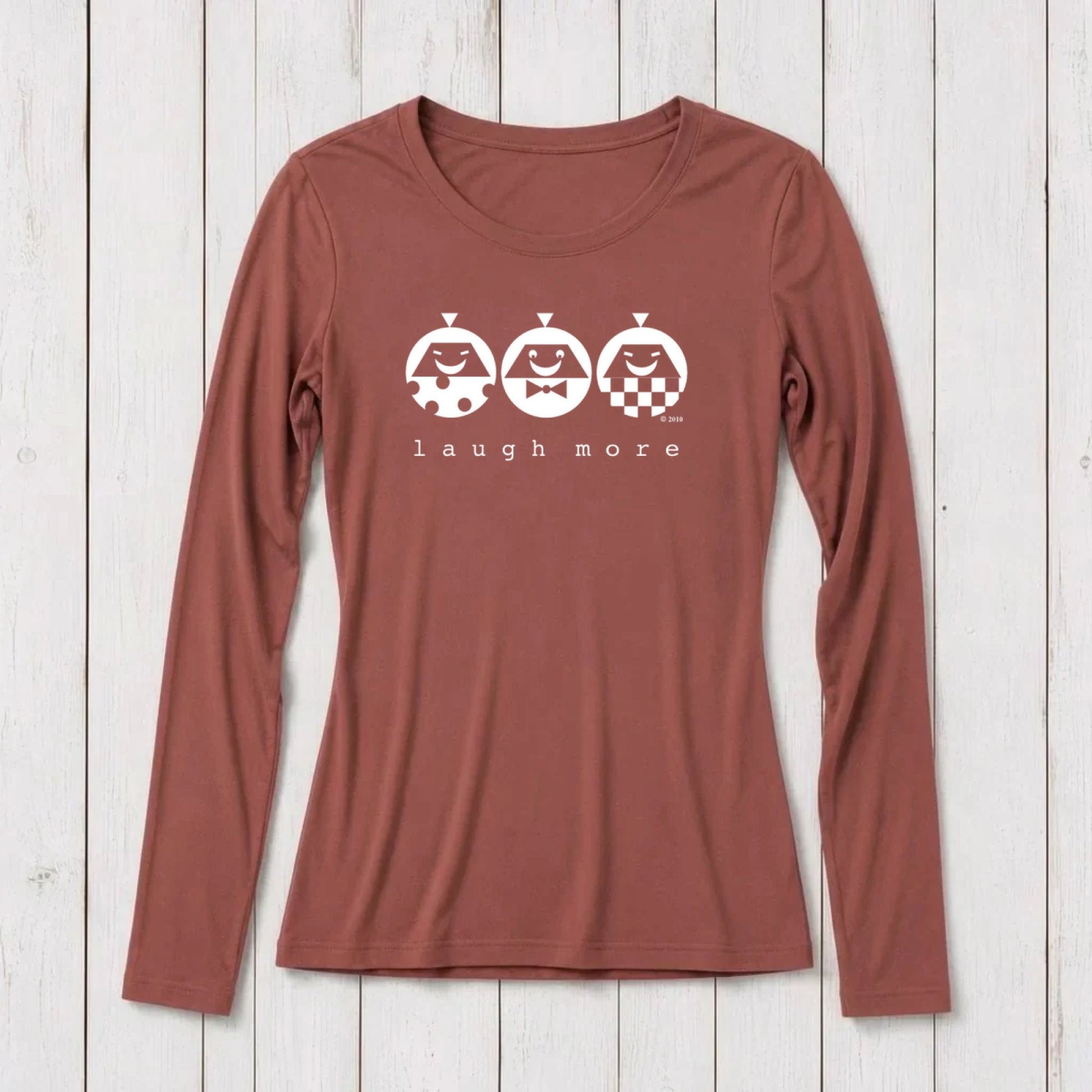 Rust-colored long sleeve T-shirt for women with white graphic design of three smiling faces with different patterns and the text 'laugh more' underneath, made from bamboo and organic cotton.
