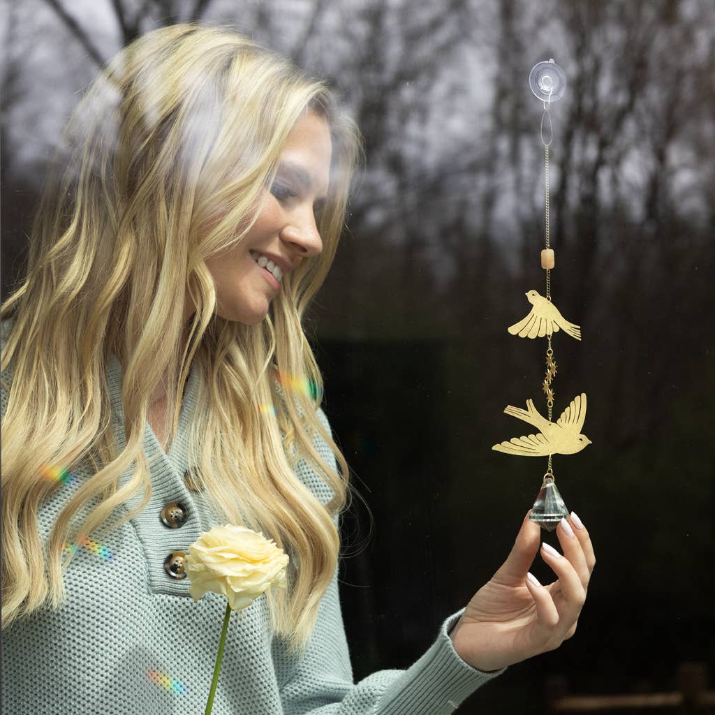 Woman holding a bird-themed suncatcher near a window, showing how sunlight creates rainbows with Scout's decorative hanging crystal.