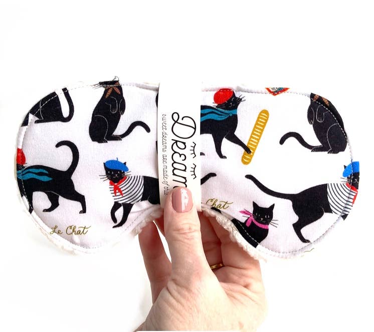 Hand holding Le Chat Cotton Sleep Mask with whimsical black cat design, made from 100% cotton and soft Sherpa for restful sleep.