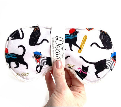 Hand holding Le Chat Cotton Sleep Mask with whimsical black cat design, made from 100% cotton and soft Sherpa for restful sleep.