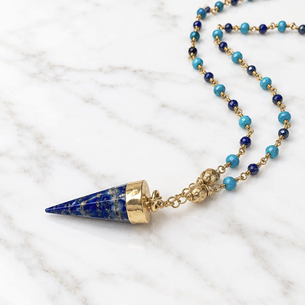 A beaded necklace featuring a pointed lapis lazuli pendant capped in gold, suspended from a chain of turquoise and dark blue beads with gold spacers, laid out on a plain off-white surface.