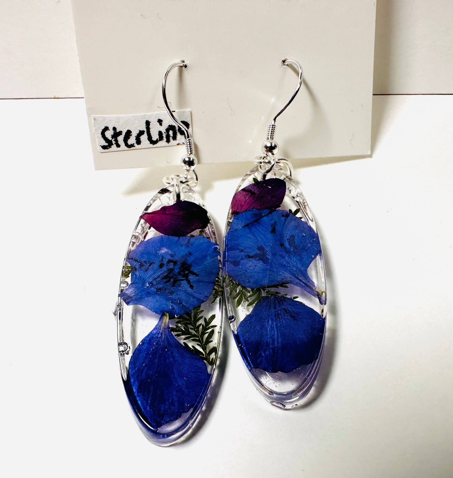 Oval resin earrings with blue and purple flowers and silver hooks. Elegant real flower earrings with a cool-toned floral design.