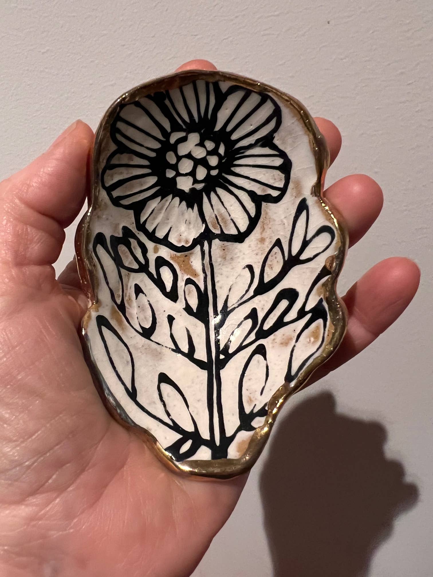 Hand holding a 5" handmade floral shaped 22K ceramic catchall jewelry dish with black flower and leaf illustration and gold edging.