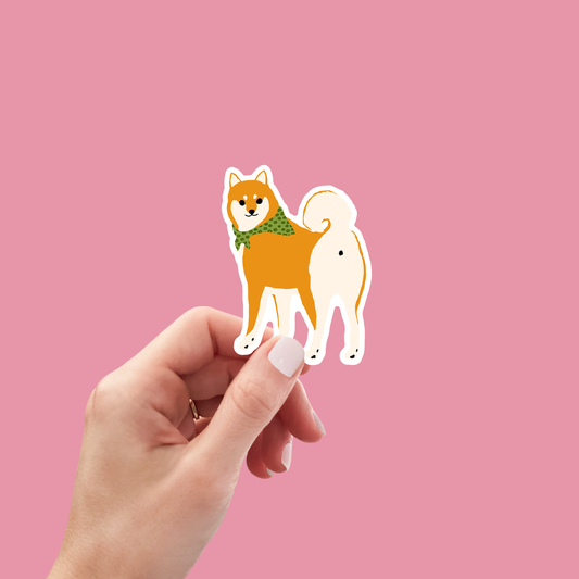 Hand holding a Shiba Inu vinyl sticker on a pink background, showcasing its compact size and durable waterproof design.