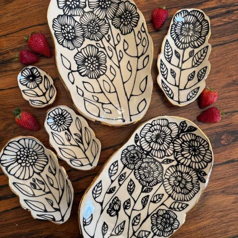 Set of 5" handmade floral shaped 22K ceramic catchall jewelry dishes with black botanical designs on cream glaze, displayed on wood with strawberries.