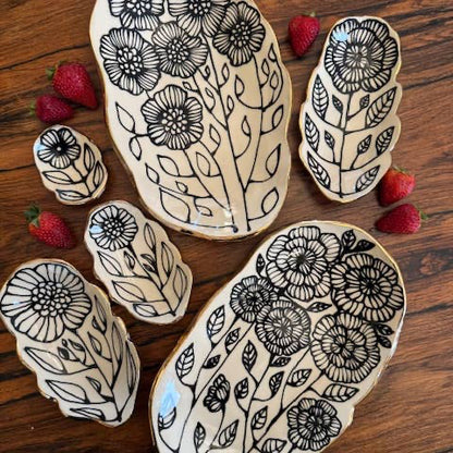 Set of 5" handmade floral shaped 22K ceramic catchall jewelry dishes with black botanical designs on cream glaze, displayed on wood with strawberries.