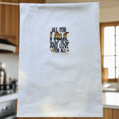 Soft flour sack kitchen towel hanging in a cozy kitchen, featuring rainbow embroidered 'All for Love and Love for All' message at the center.