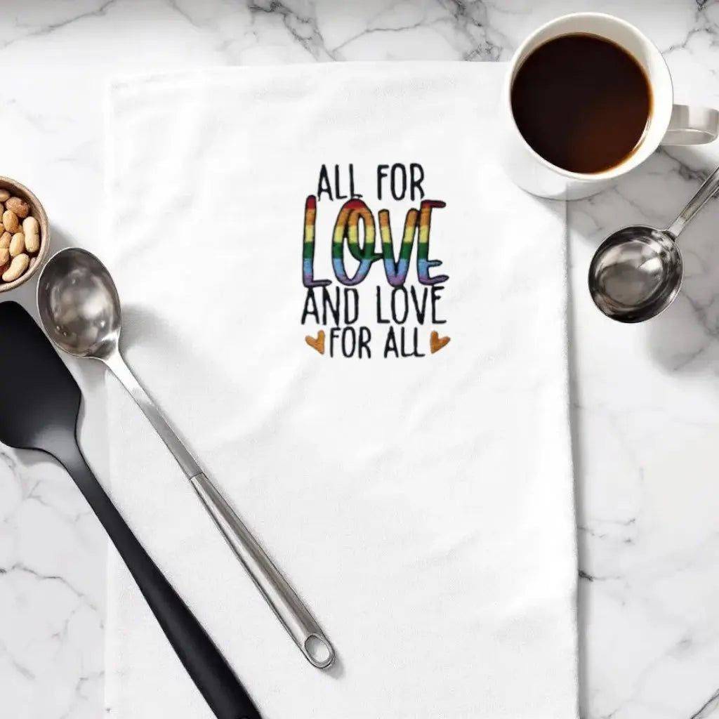 Embroidered kitchen towel with 'All for Love and Love for All' text in rainbow colors, placed beside coffee and utensils on a marble counter.