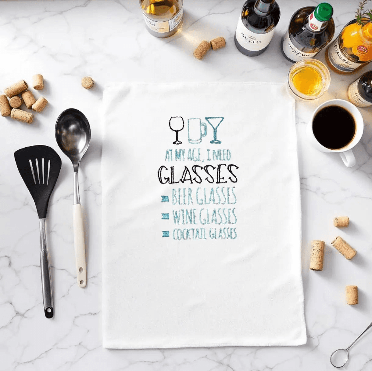 Embroidered towel with humorous 'At my age I need glasses' phrase, surrounded by kitchen utensils and wine bottles. Ideal funny kitchen gift.