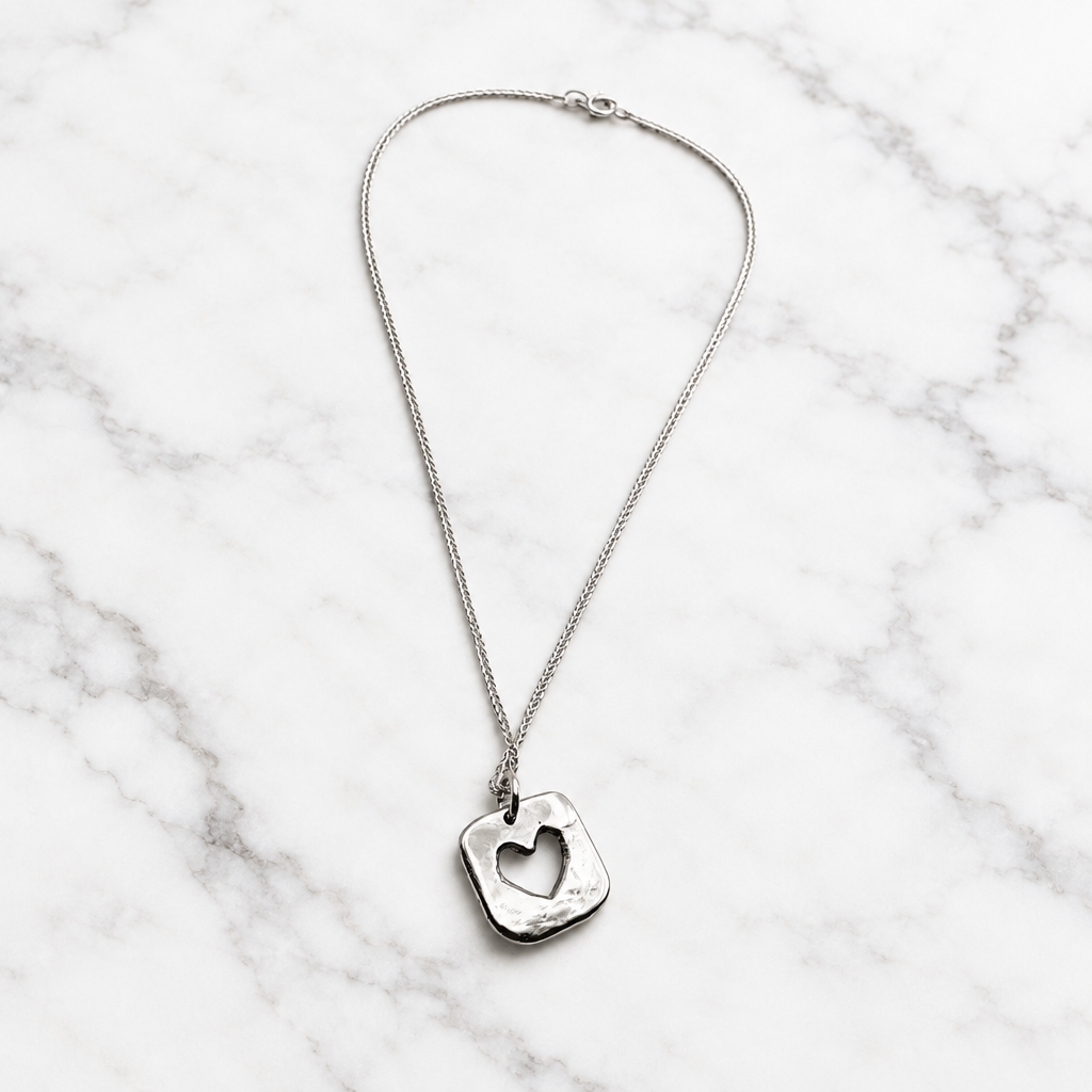 A silver pendant necklace featuring a square, openwork charm with a heart cutout in the center, suspended from a delicate silver chain against a plain white background.