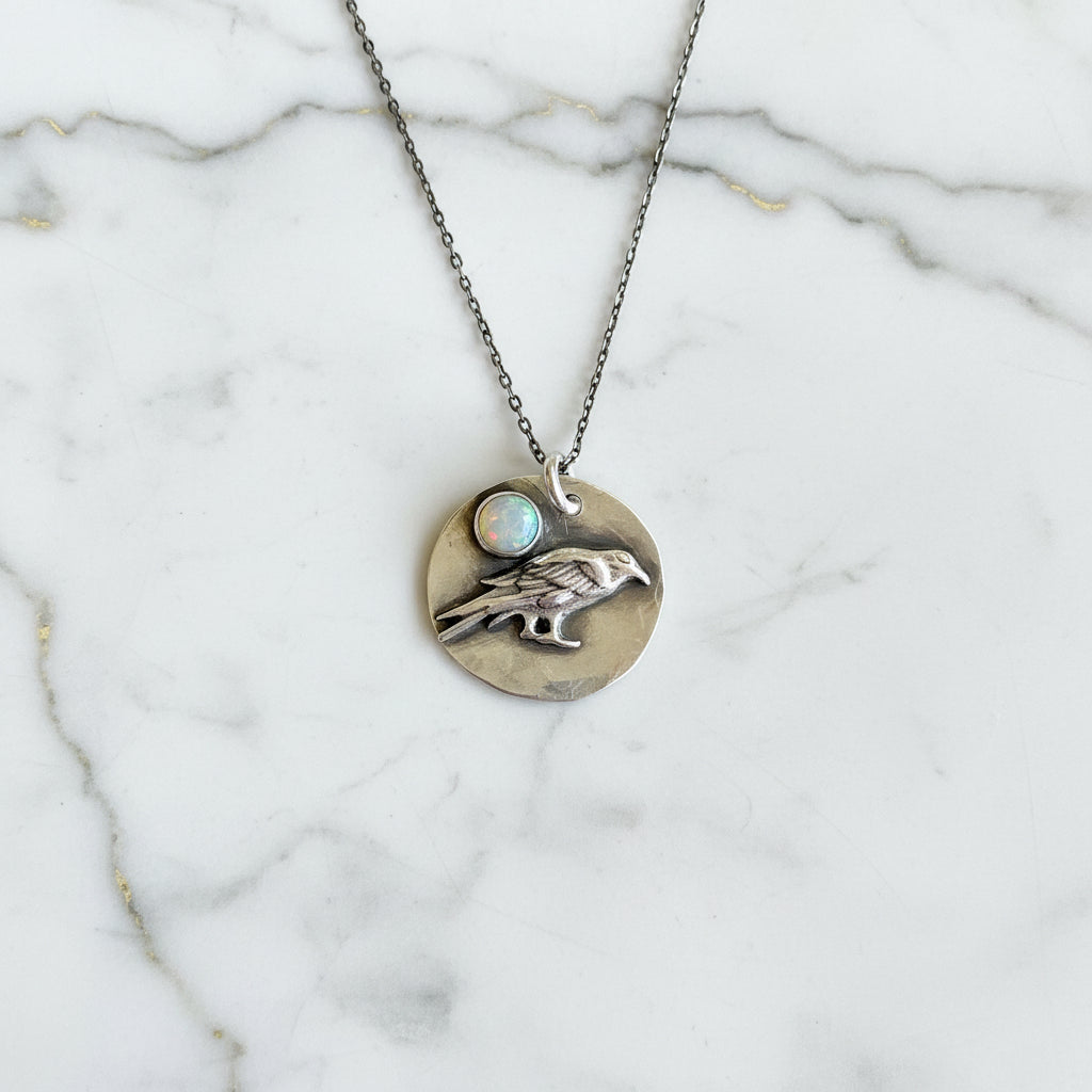 Silver necklace with a round pendant featuring a hand and opal stone on a textured gray surface.