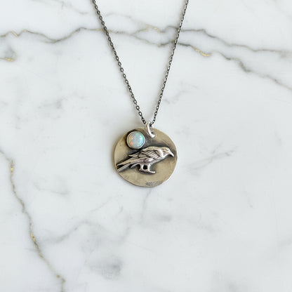 Silver necklace with a round pendant featuring a hand and opal stone on a textured gray surface.