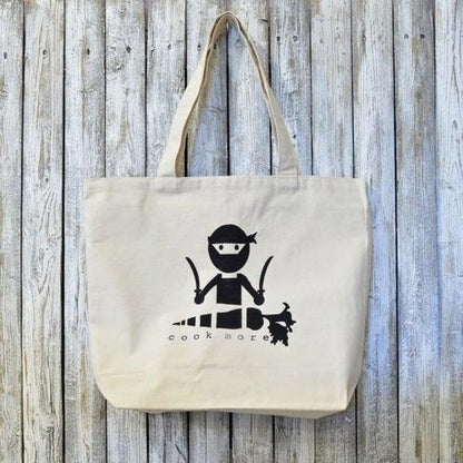 Culinary tote bag organic with COOK MORE artist design featuring a ninja chef graphic, made from 100% cotton. Ideal for eco-conscious cooking lovers.