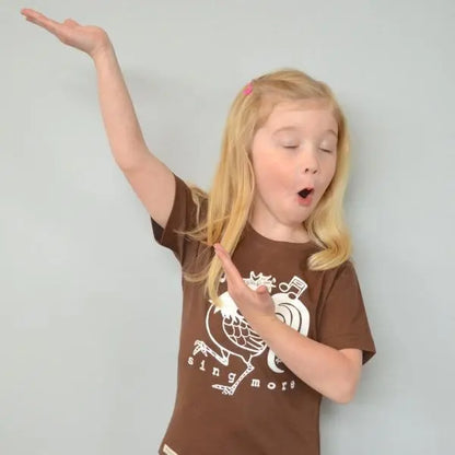Child playfully posing in brown rooster graphic tee with 'Sing More' text, organic cotton shirt for kids with fun animal design.