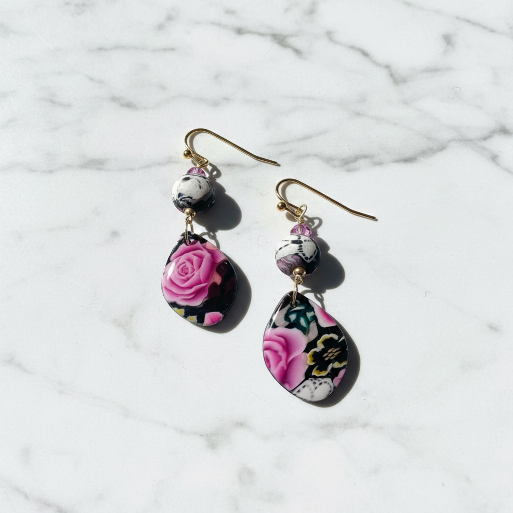 A pair of dangle earrings with gold-tone fishhook wires, small marbled beads, and teardrop-shaped charms featuring a pink rose and floral pattern on a black background, set against a plain white background.