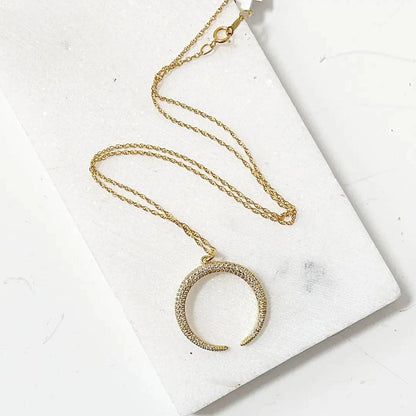 Gold crescent moon necklace with cubic zirconia on an 18” rope chain, displayed on a white marble background. Elegant and ideal for layering.