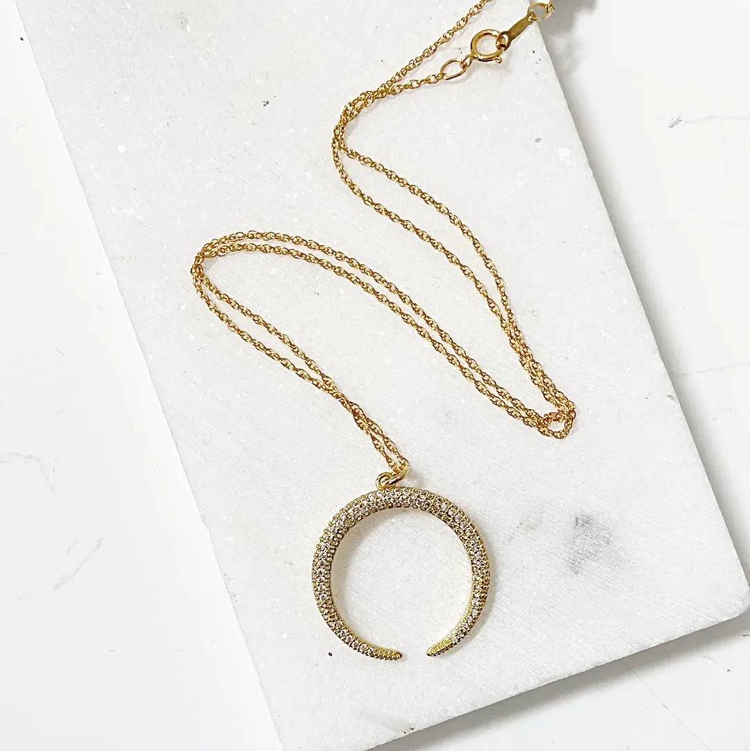 Dainty gold crescent moon necklace with sparkling cubic zirconia on an 18” rope chain, laid flat on a white surface for product showcase.