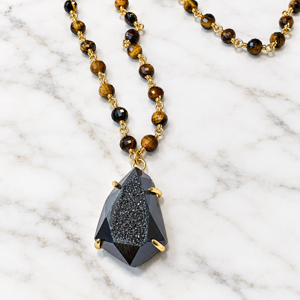 A beaded necklace featuring round brown and black tiger's eye beads with gold spacer rings, hanging from a gold-toned chain with a faceted dark gray drusy gemstone pendant, displayed on a plain off-white surface.