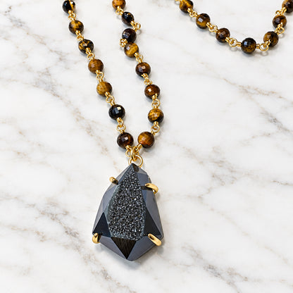 A beaded necklace featuring round brown and black tiger's eye beads with gold spacer rings, hanging from a gold-toned chain with a faceted dark gray drusy gemstone pendant, displayed on a plain off-white surface.
