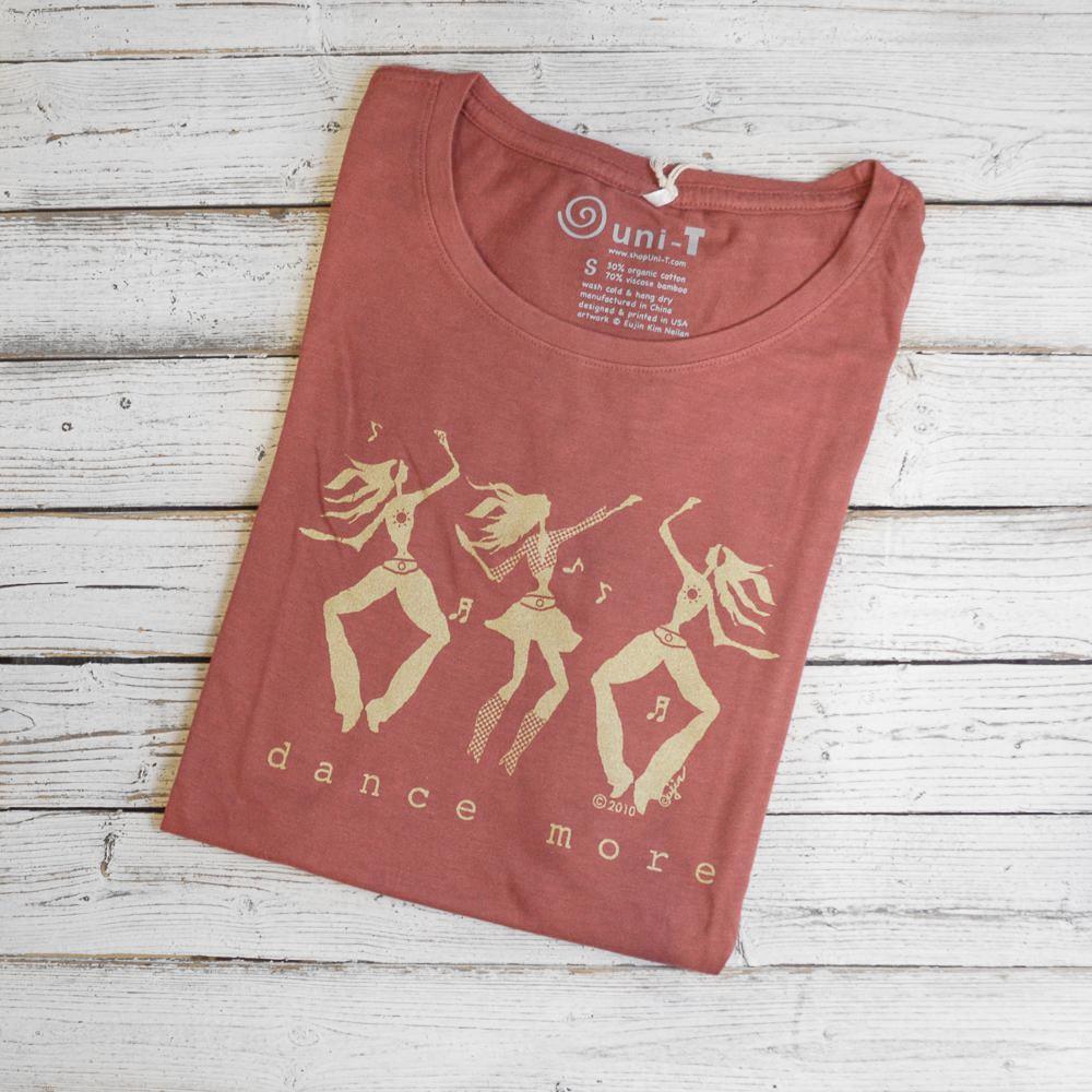 Women's Dance T-Shirt in Bamboo & Organic Cotton in rose color with 'Dance More' dancer graphic folded on wood background. Ideal gift for dancers.