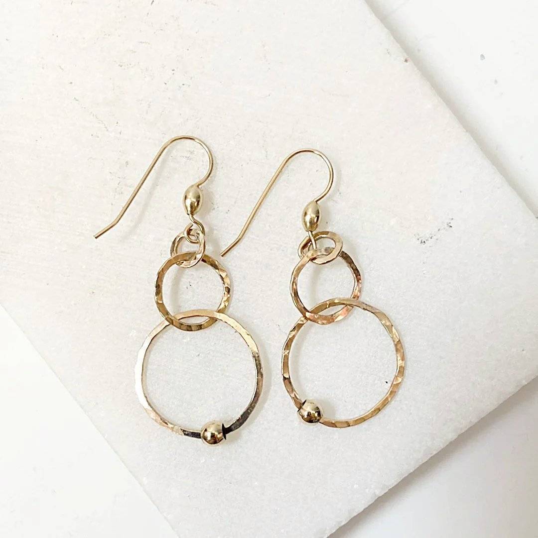 Pair of gold HoopLink Earrings with interlinked hammered hoops and bead detail, laid flat on a white stone surface for product display.
